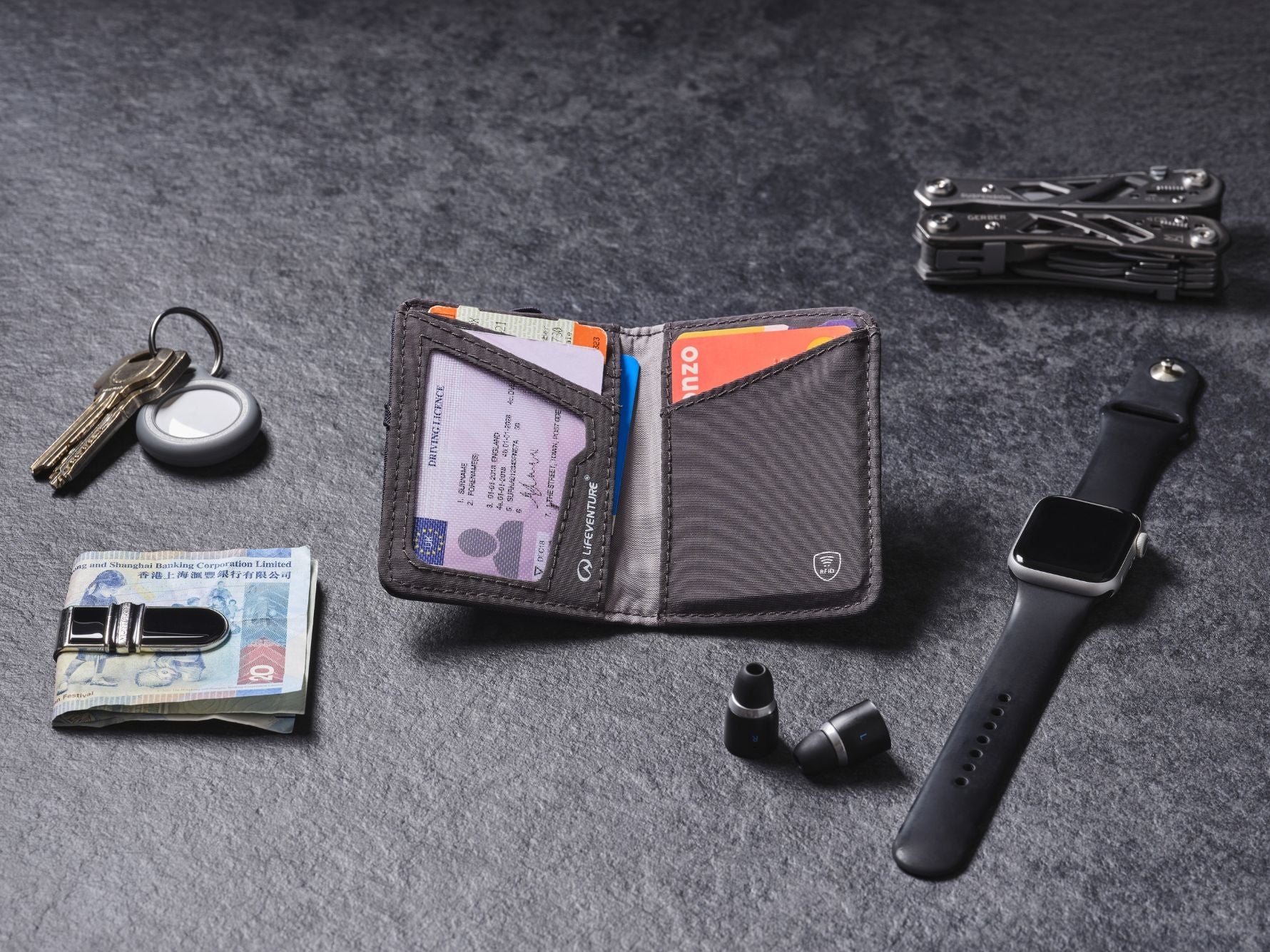 Open Lifeventure RFID wallet with cards, cash and passport laid out alongside everyday travel essentials on a dark surface.