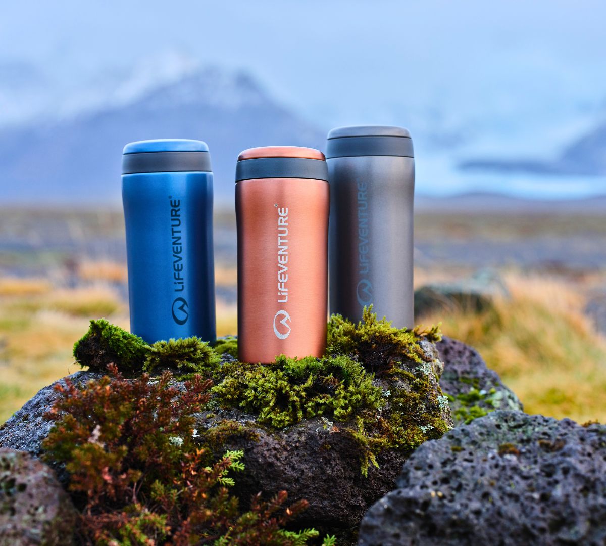 Three Lifeventure Thermal Mugs in Blue, Copper, and Grey, placed on mossy rocks with a blurred mountain landscape in the background.