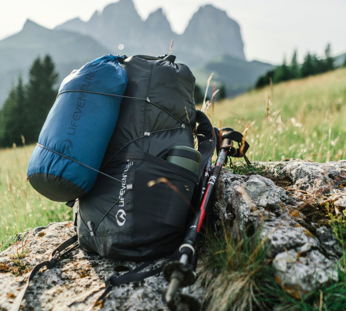 A Lifeventure packable backpack with attached blue sleeping bag and hiking poles resting on a rocky outcrop in a grassy mountain landscape.