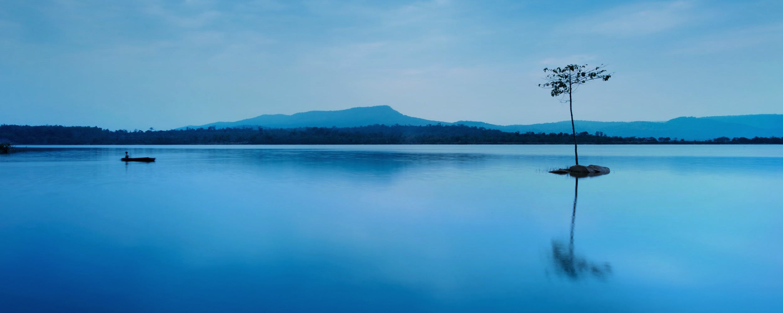 A lone tree stands on a still lake with its reflection mirrored in the water. A small boat floats nearby, surrounded by distant mountains.