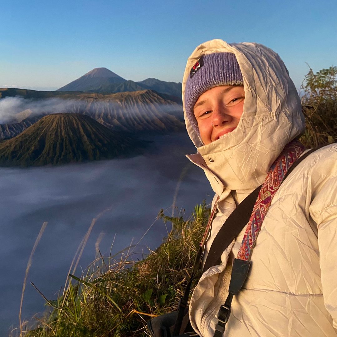 Emily dressed in a warm jacket, hat, and scarf smiles at the camera with a misty mountain landscape and volcano in the background.