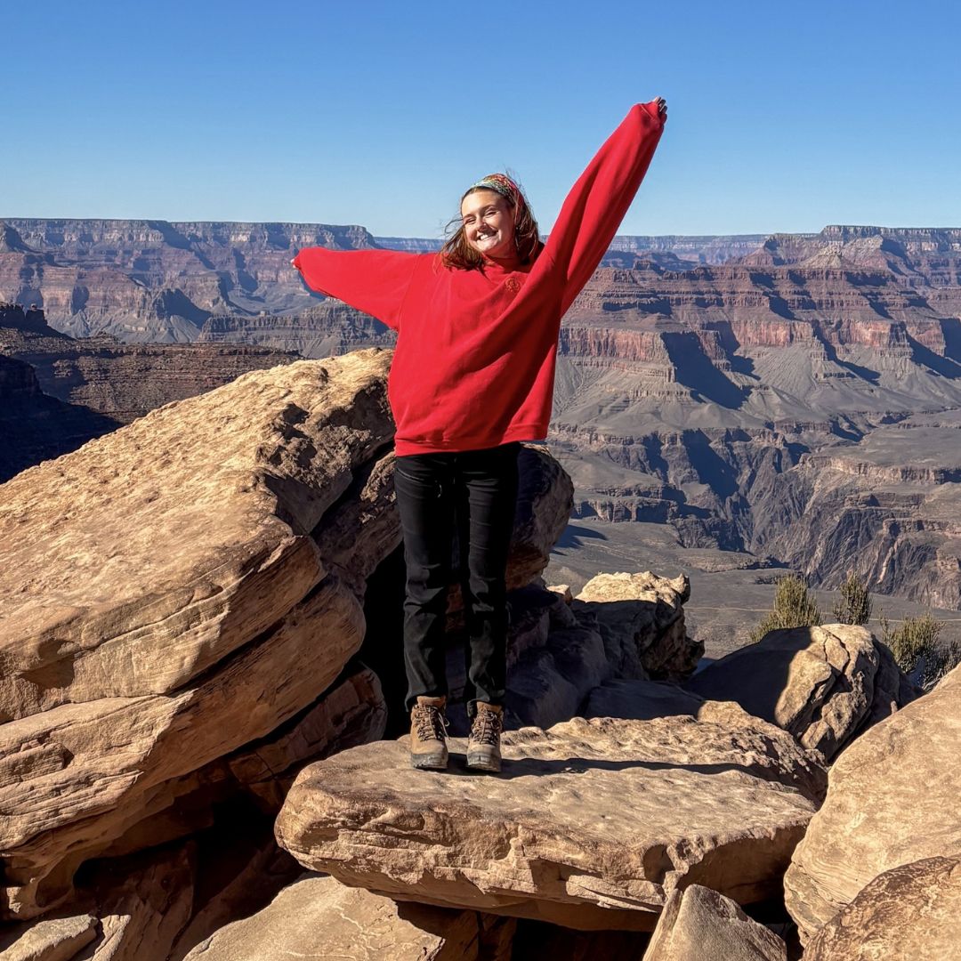 Emily wearing a red jumper and hiking boots stands triumphantly on rocky cliffs with arms raised, overlooking a vast canyon landscape.