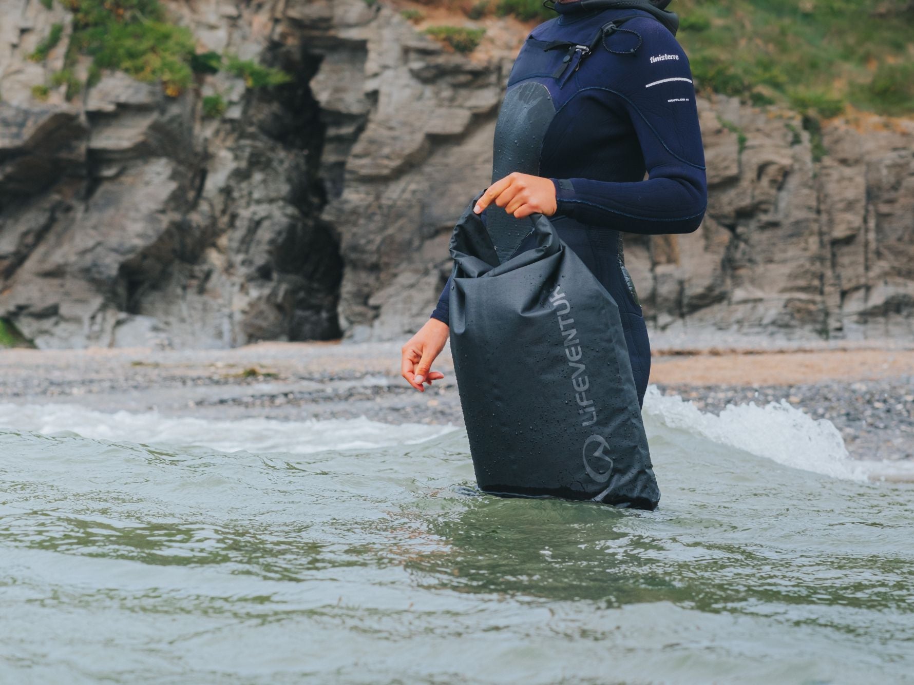 Person standing in the sea holding a black Lifeventure Storm Dry Bag, demonstrating its heavy-duty waterproof performance in coastal conditions.