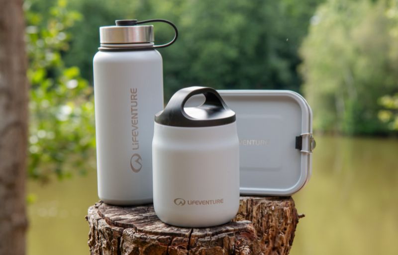 Lifeventure insulated Food Flask, Stainless Steel Flask and Lunch Box in white, displayed on a tree stump beside a lake with a forest backdrop.