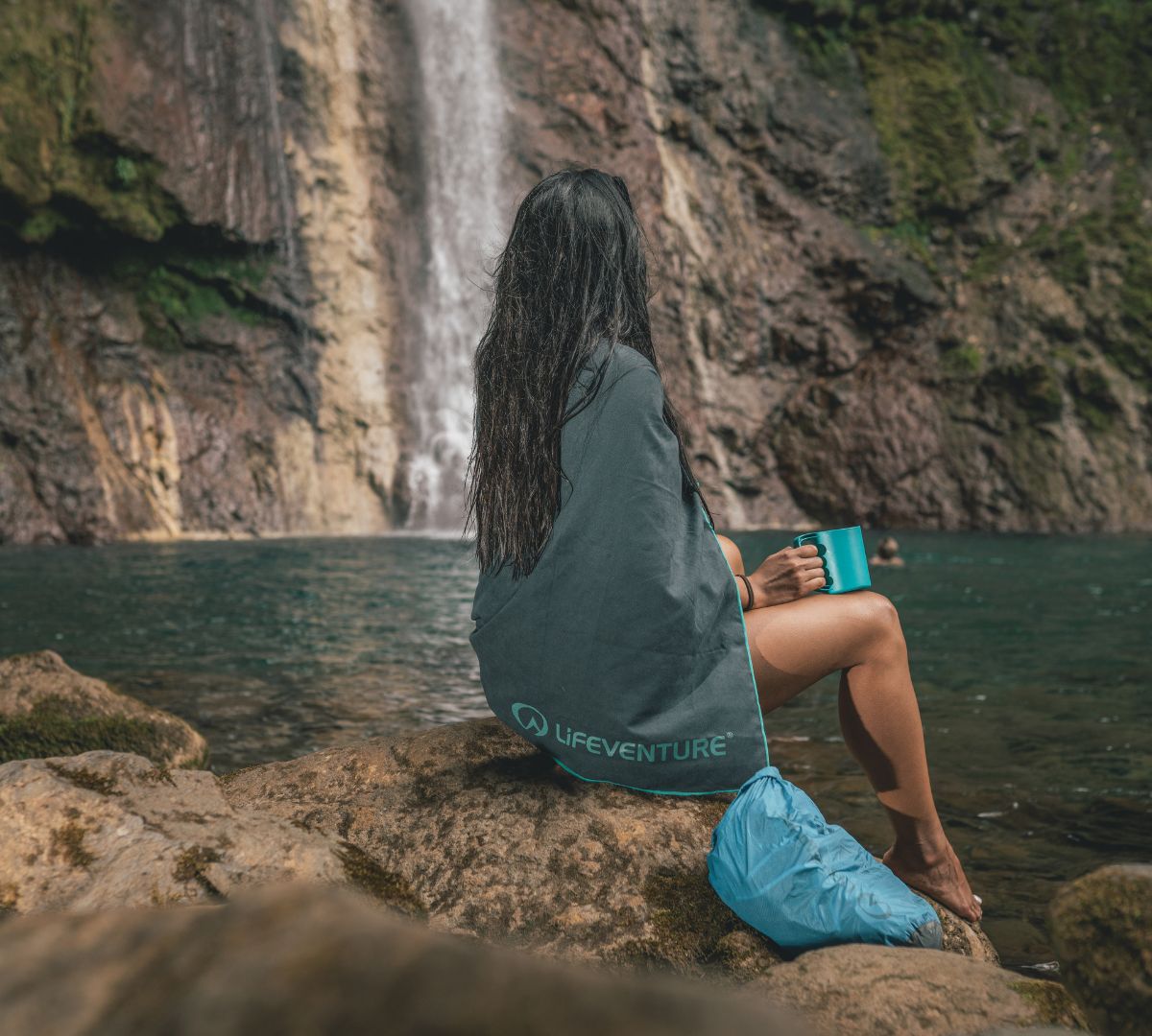 Woman sitting by a waterfall wrapped in a grey Lifeventure towel featuring Polygiene StayFresh technology, holding a blue mug.