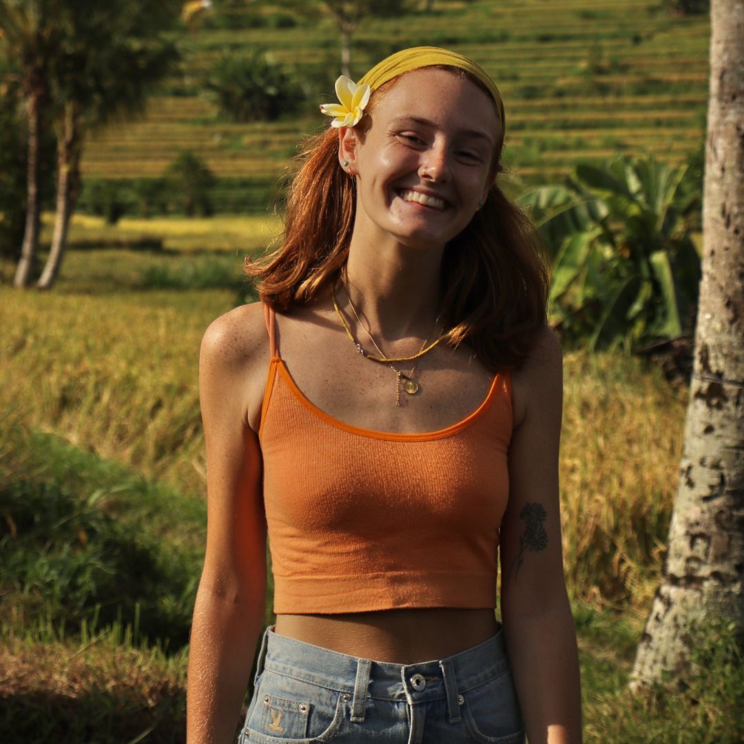 A smiling Emily with a yellow headband and flower in her hair stands in a sunlit rice field, wearing an orange crop top and denim jeans.